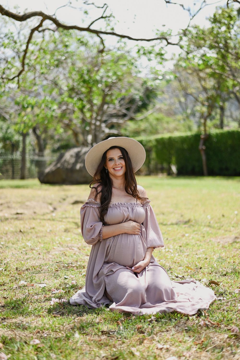 Expectant mother in off-shoulder mauve dress sits gracefully on grass in outdoor garden setting.