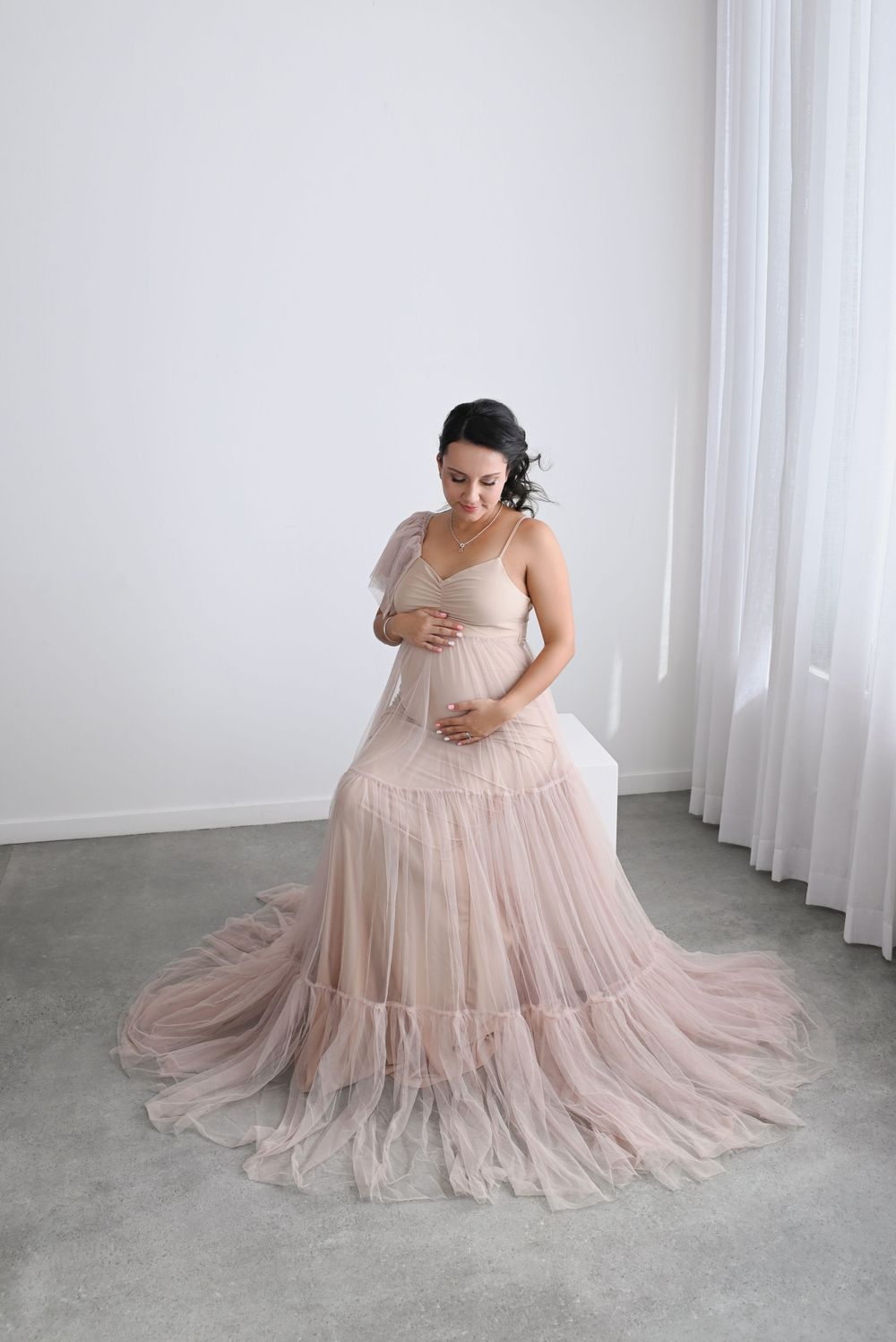 Elegant maternity portrait in sweeping blush gown with tiered tulle skirt on concrete floor.