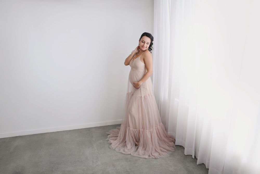 Side view maternity pose in romantic blush tulle gown against minimalist white background.