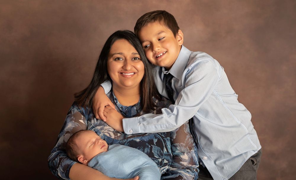 A family portrait featuring two siblings and a newborn baby against a brown backdrop.