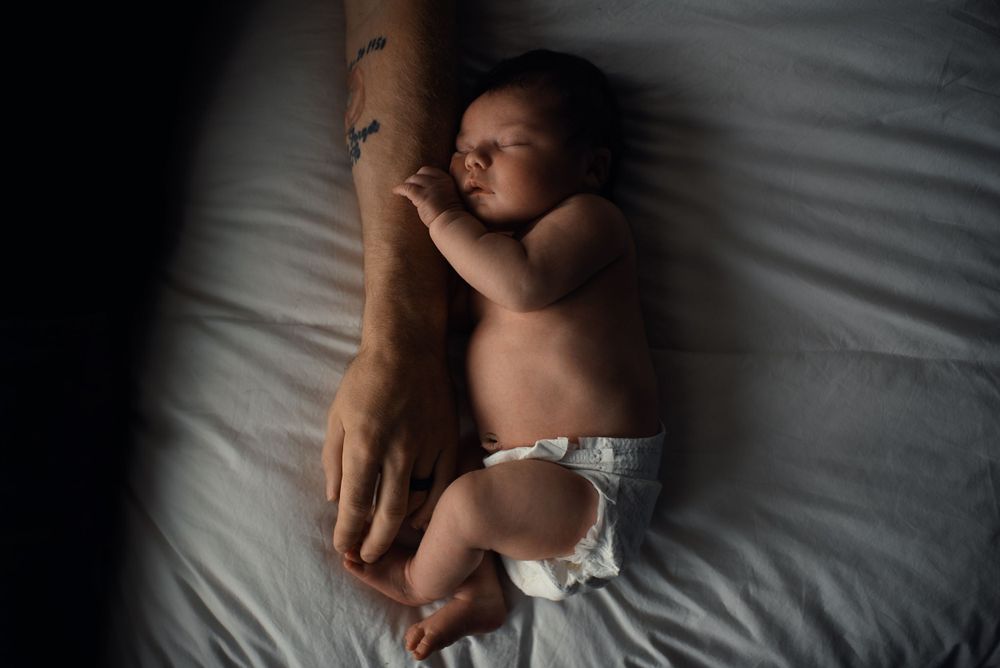 A newborn baby sleeps peacefully on white bedding in natural lighting.