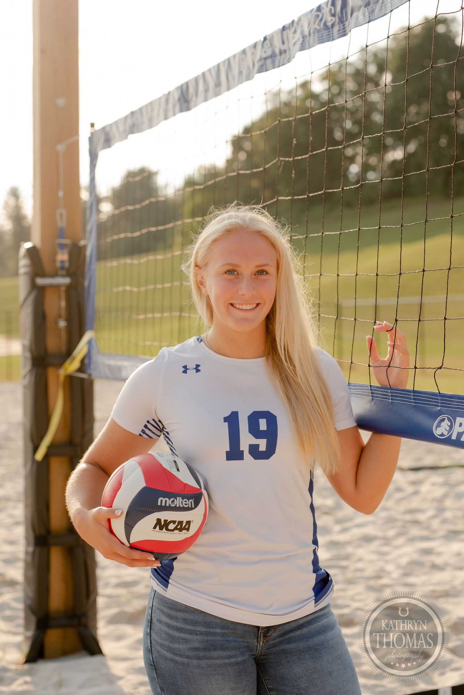 A volleyball player in jersey 19 poses by the net holding an NCAA regulation ball.