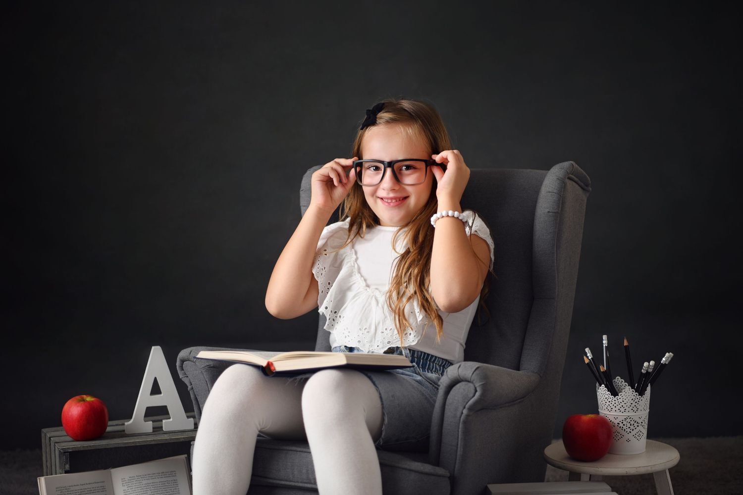 A student sits in a grey armchair with an open book and red apples, wearing glasses against a dark background.