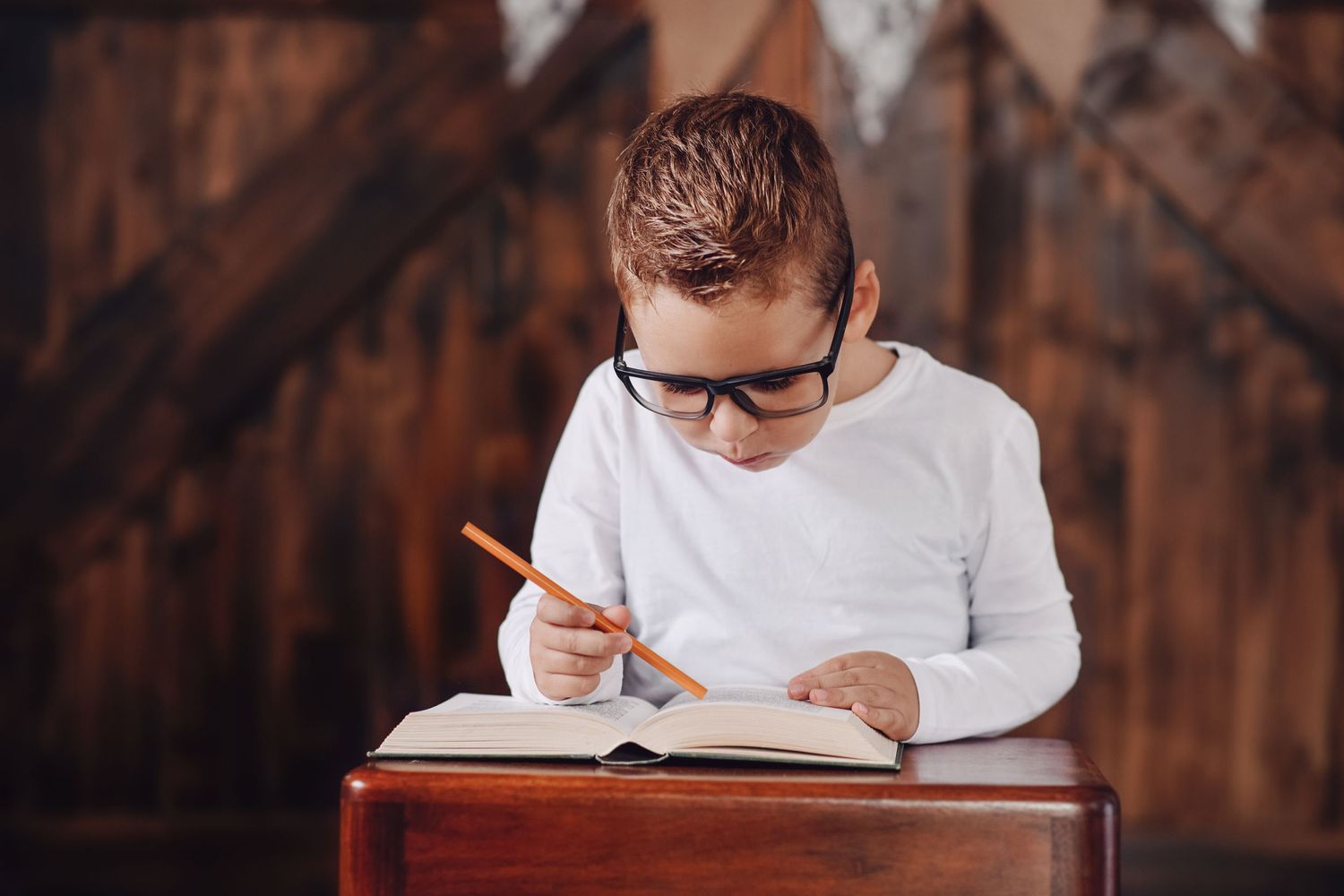 Student in glasses studies and writes in notebook at wooden desk.