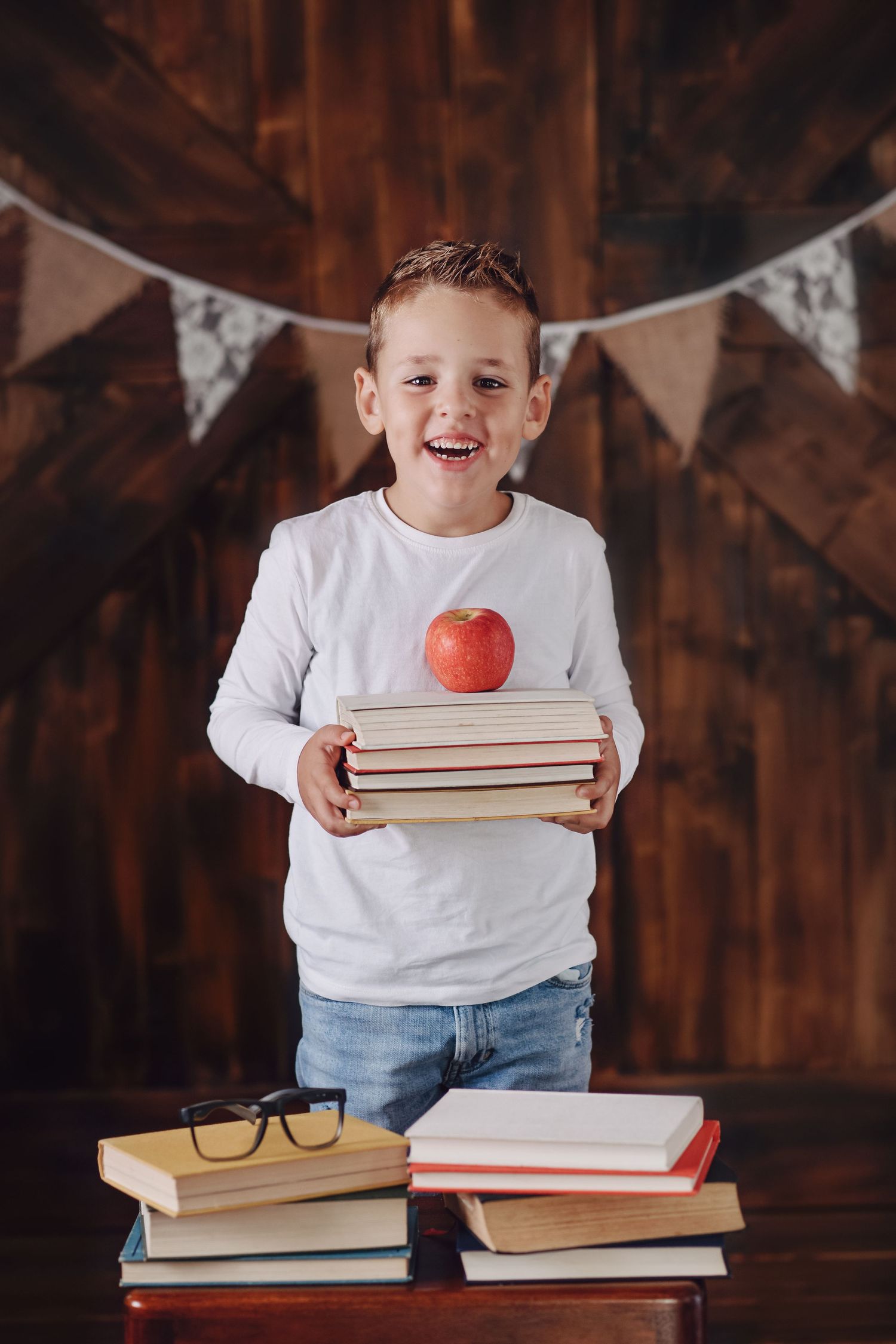 Student holds stack of books and red apple against rustic wood wall with bunting decorations.