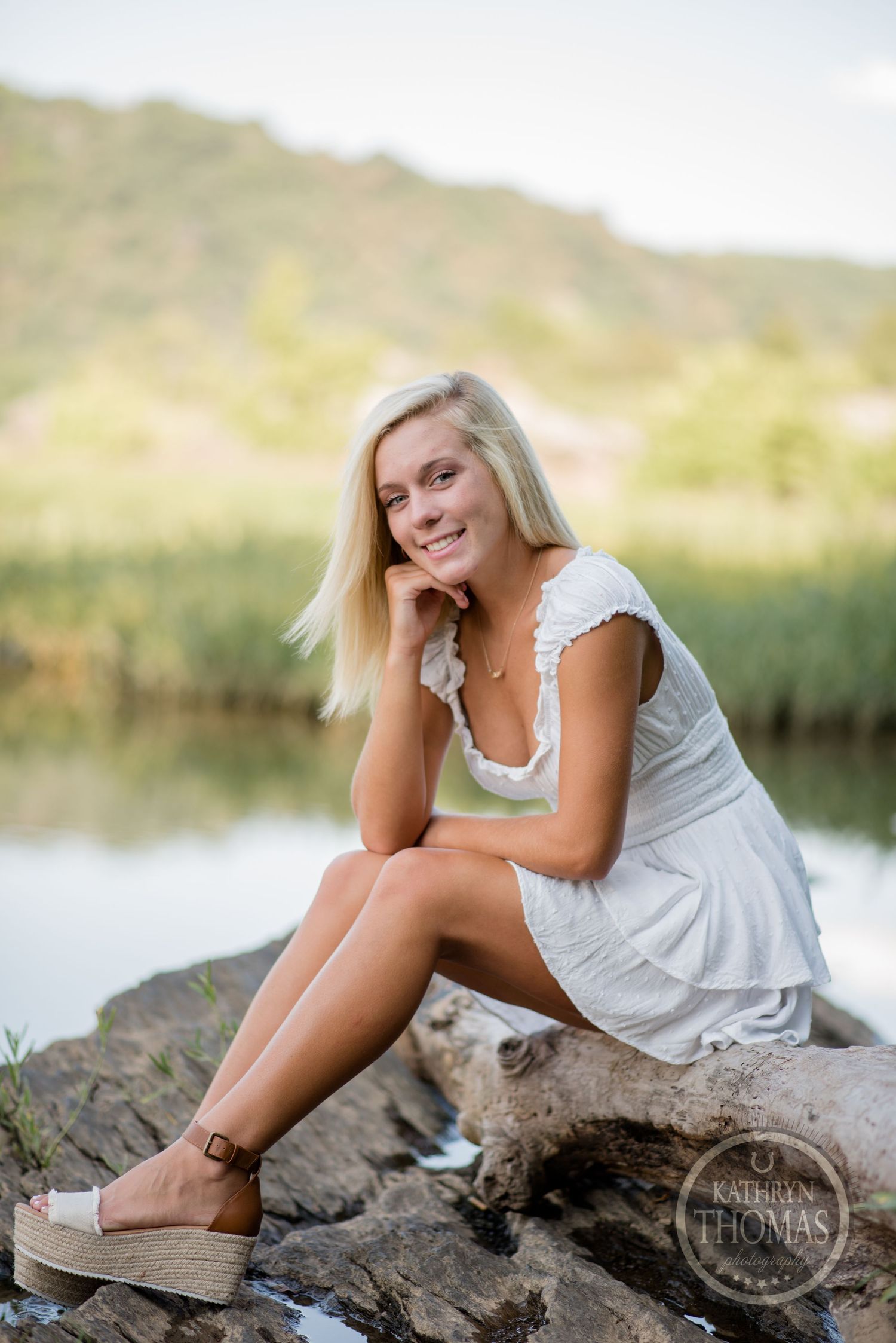 Woman in white dress posing on rocky outcrop by tranquil water.