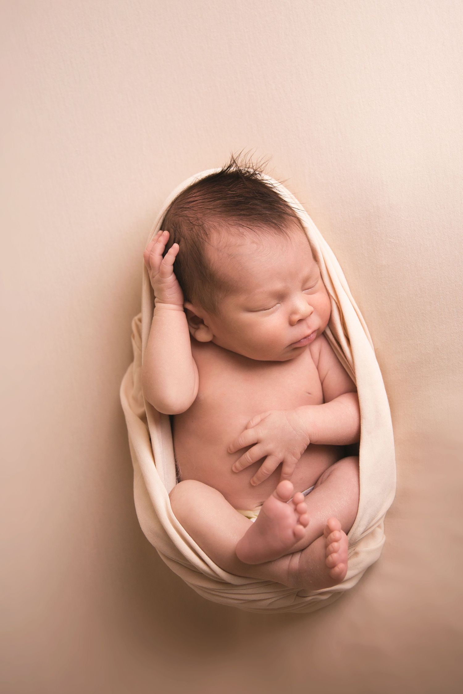 Artistic newborn photography with baby curled up in cream-colored wrap.