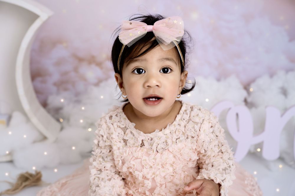 Baby in pink lace dress sits on white backdrop with dreamy star decorations for cake smash photoshoot.