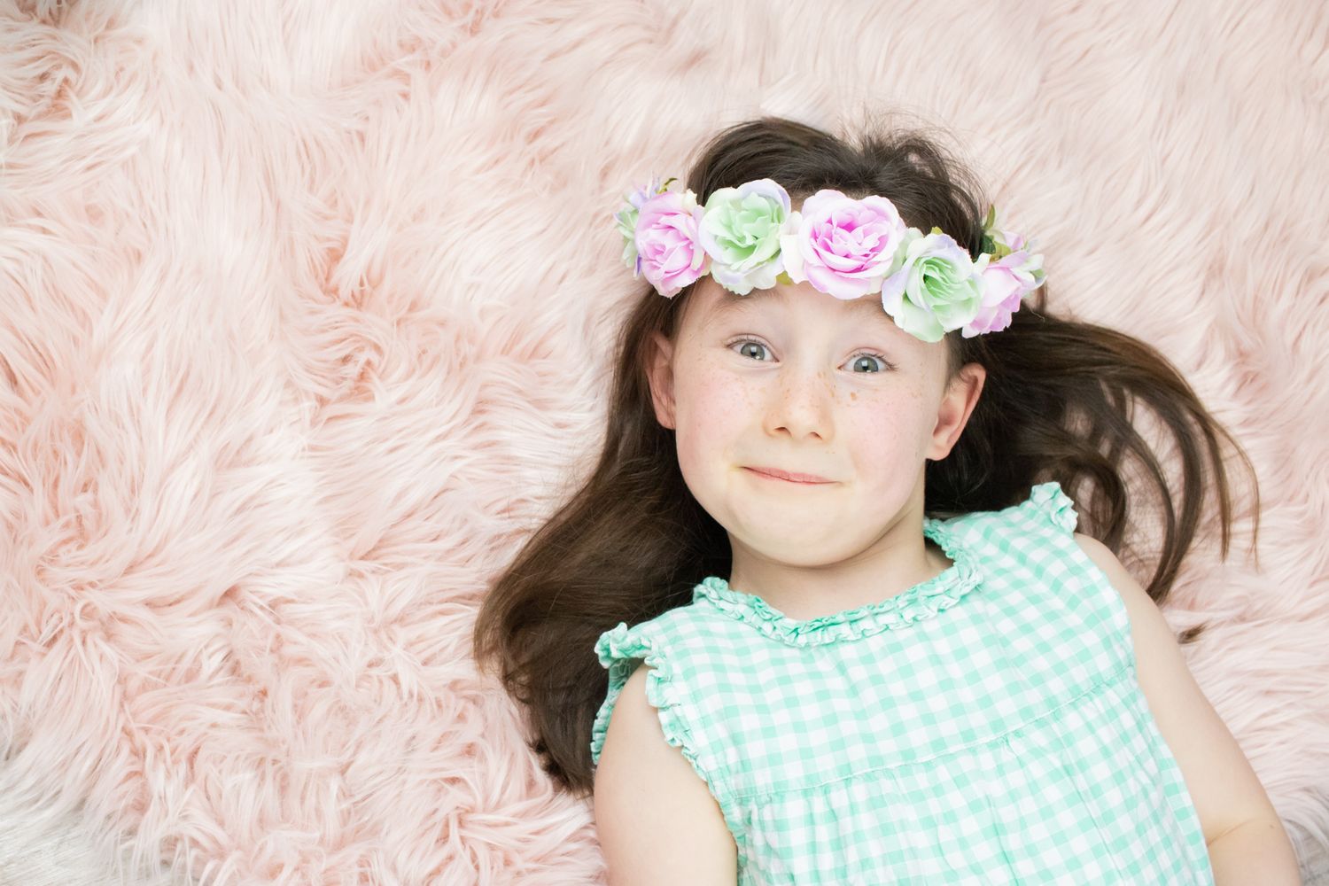 child with brown hair laid on a pink fluffy rug with flowers in her hair for a photo shoot