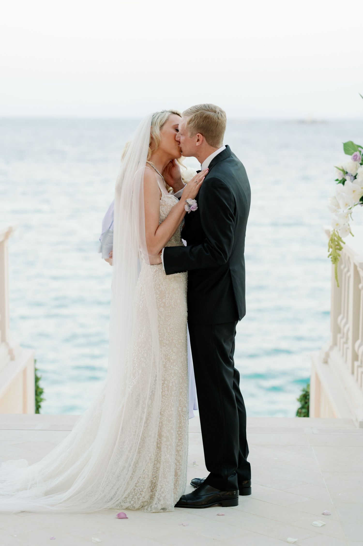 Newlyweds share first kiss at oceanfront wedding ceremony with white floral arch.