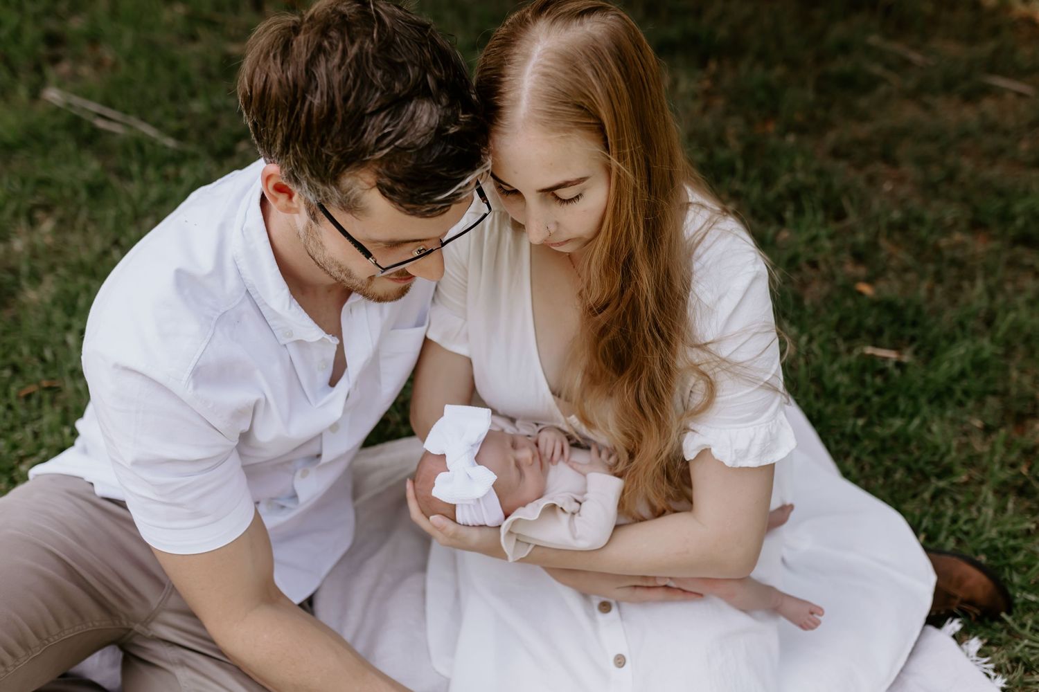 Parents in white attire sit on grass while holding their infant during an outdoor photoshoot.