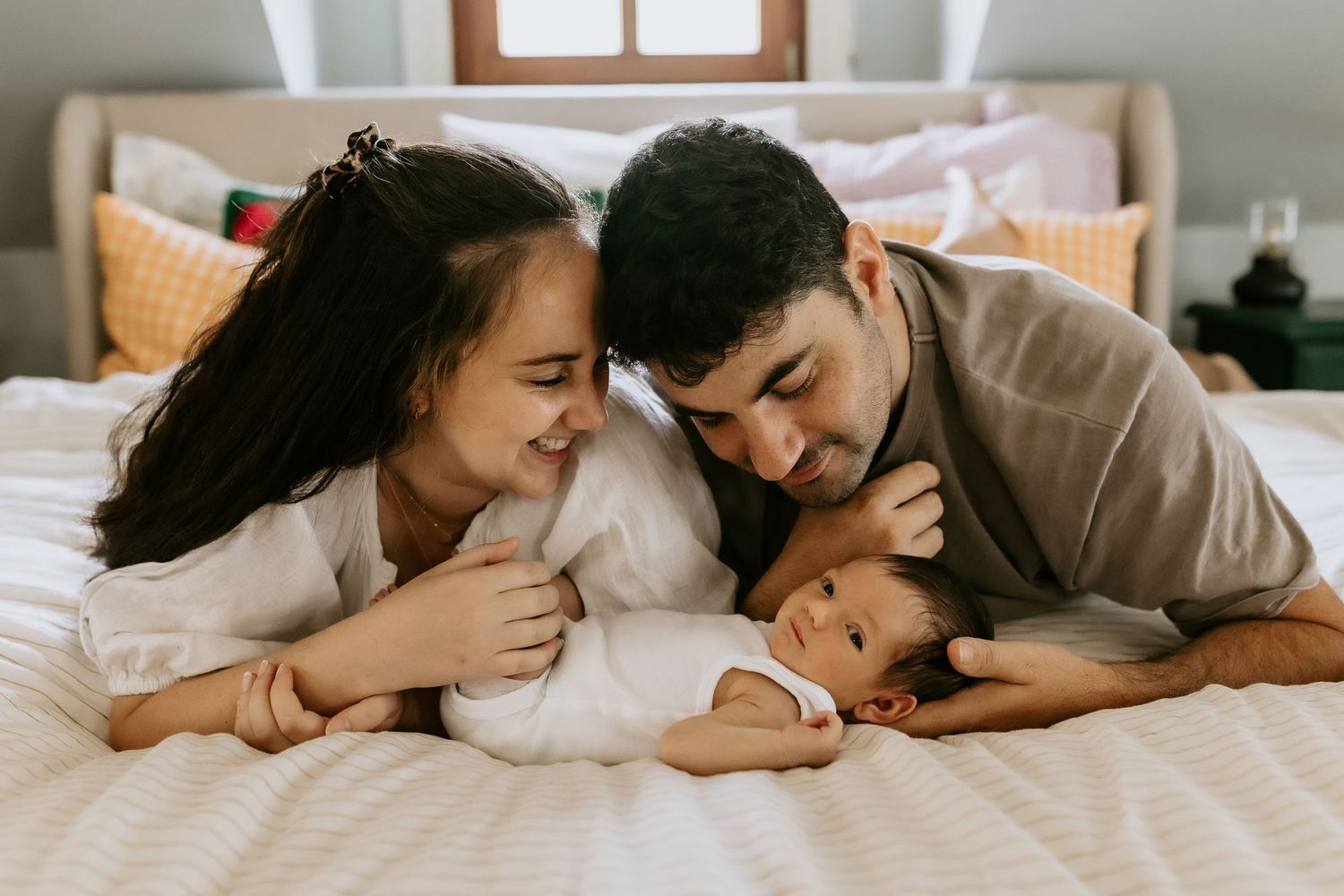Parents share intimate moments with their baby while lying on white bedding.