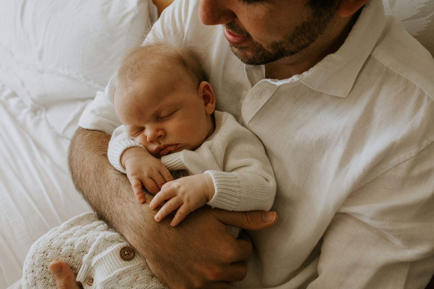 Baby in white clothing rests peacefully while parents share tender moment on bed.