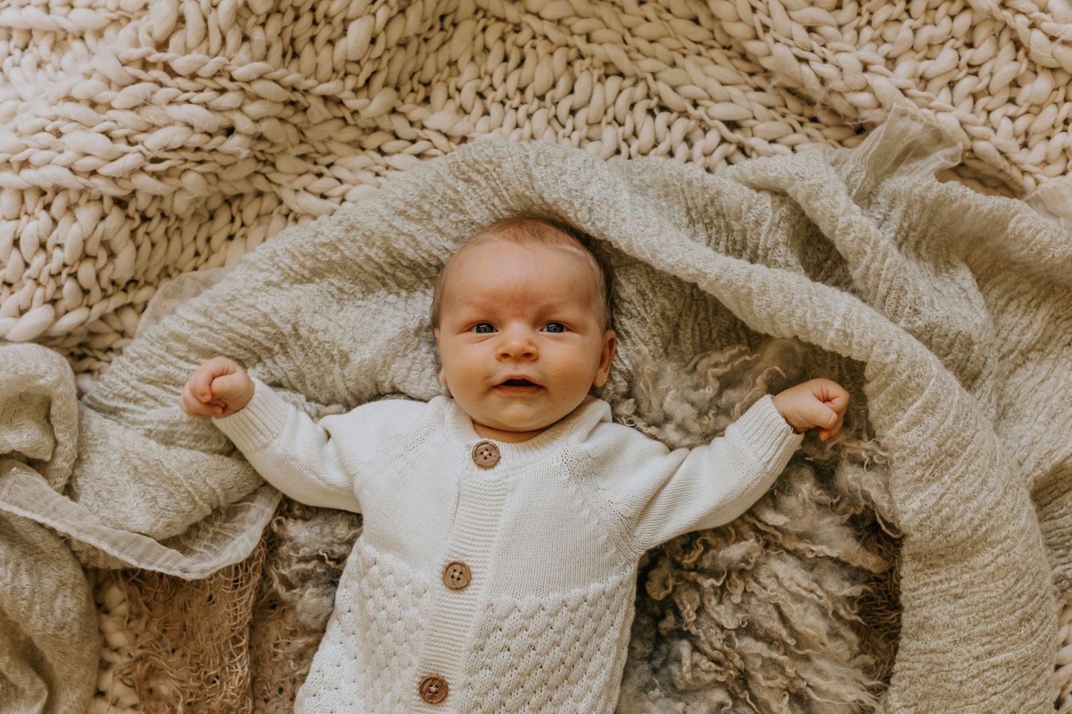 Baby in white knit outfit rests on textured beige blanket during family photo session.