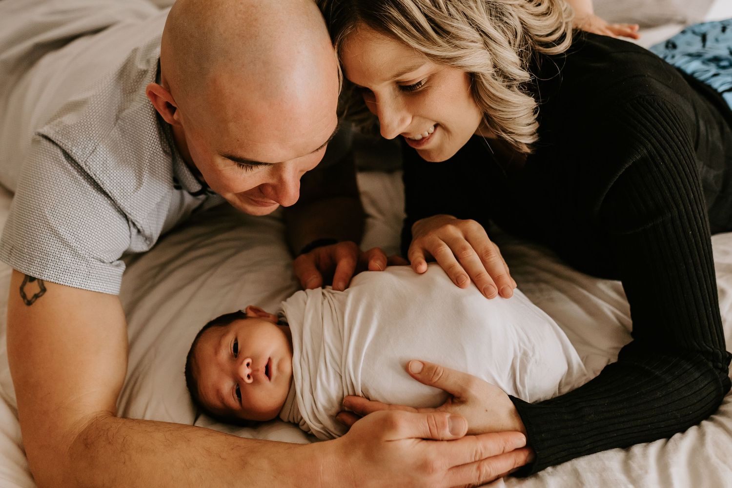 Parents lying on white bedding share intimate moments with their newborn baby during a lifestyle photography session.