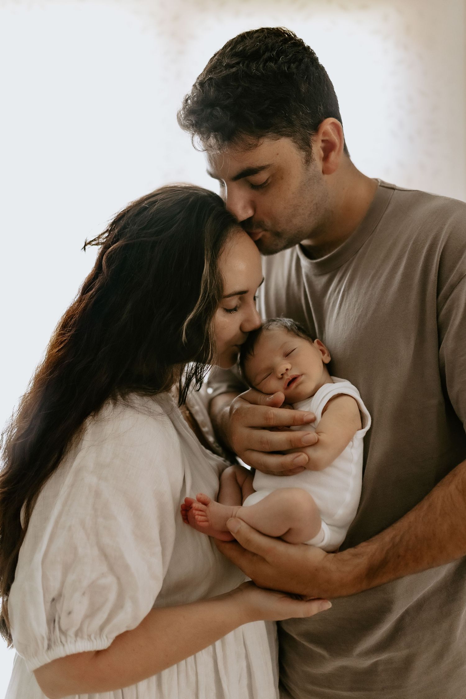 Parents holding and kissing newborn baby in intimate indoor family photoshoot.