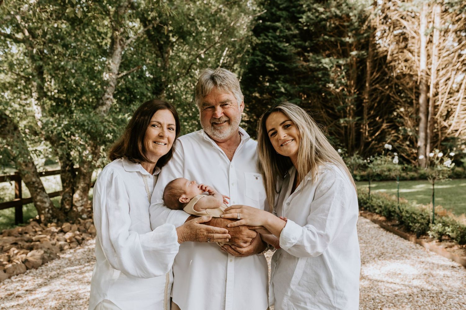 A tender family moment captured outdoors in white clothing with natural lighting and greenery in the background.