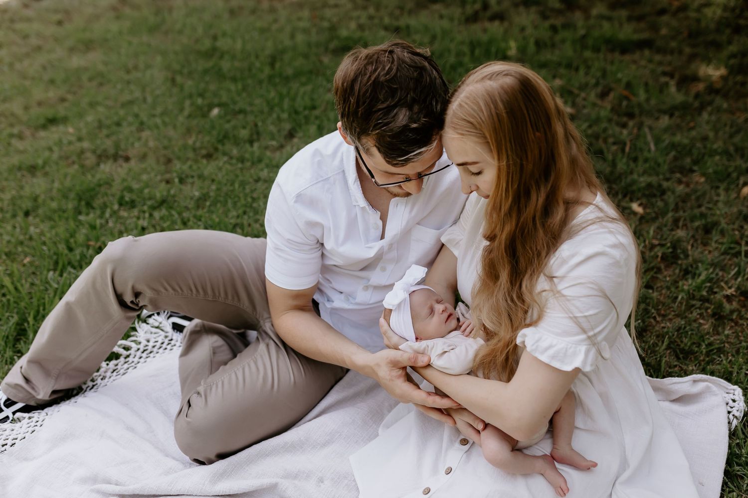 Parents in casual attire lounge on grass during outdoor family photoshoot.