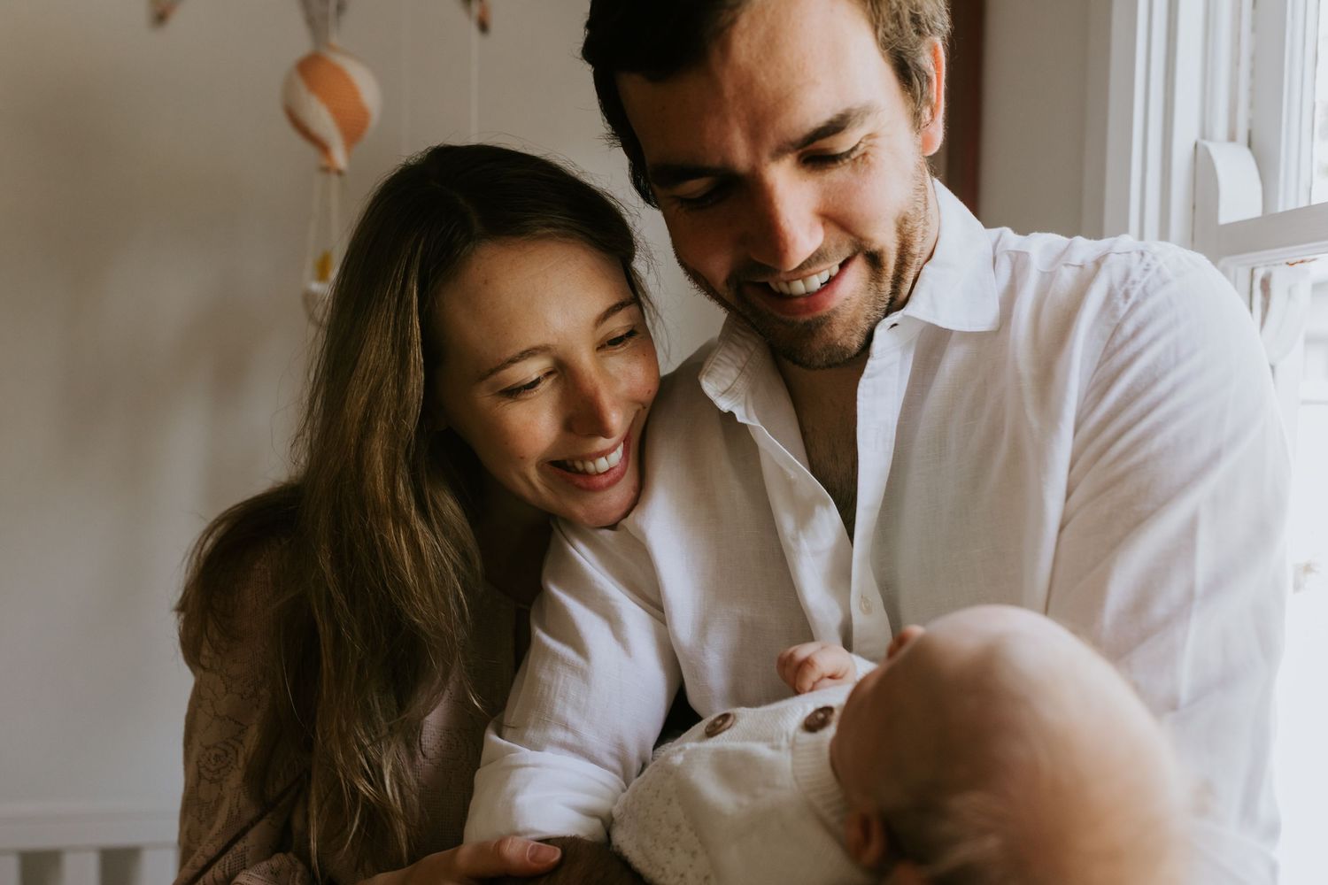 Tender family moment in natural lighting showing parents bonding with their newborn baby in white clothing.