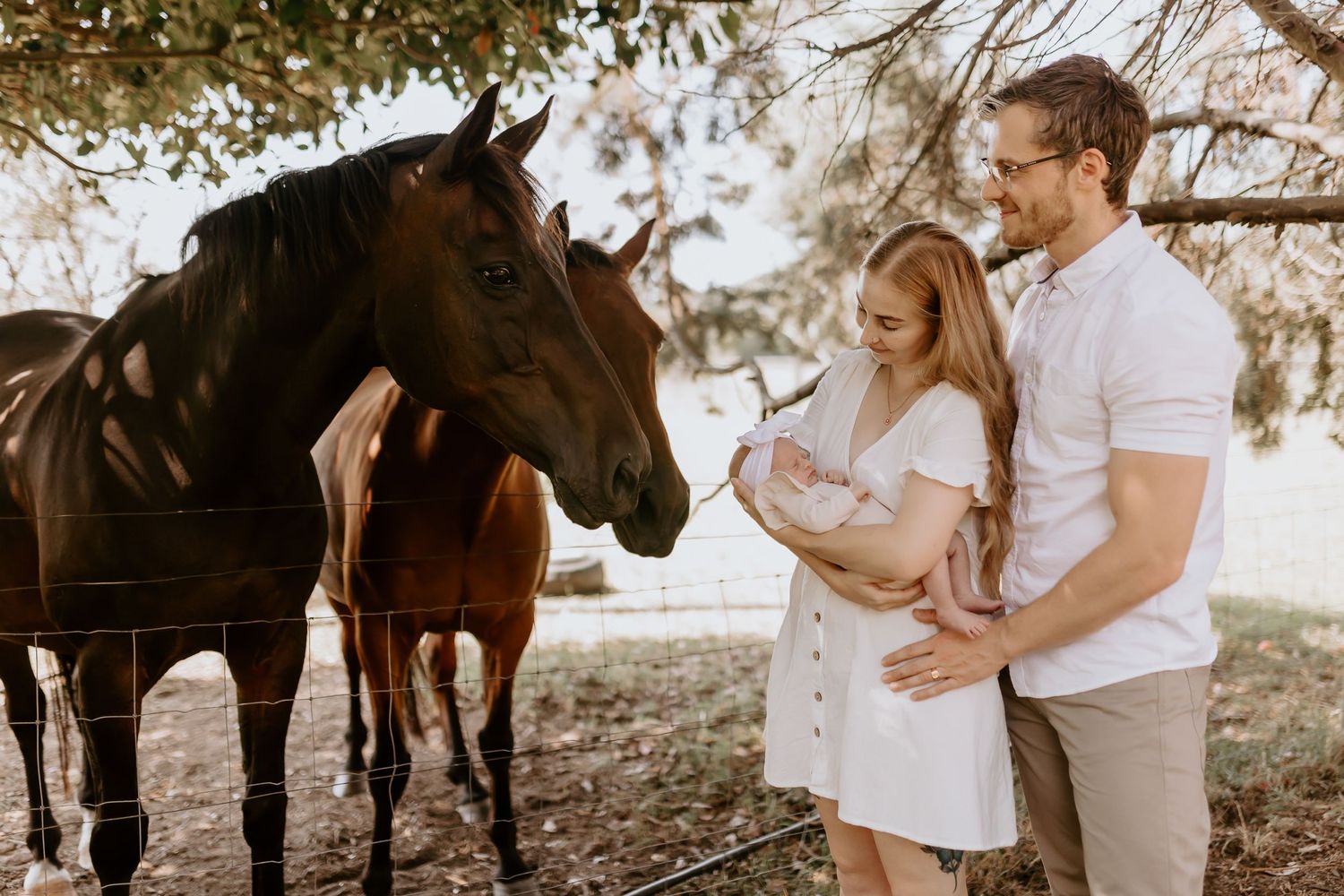 A couple in white poses with their baby outdoors with horses in the background.