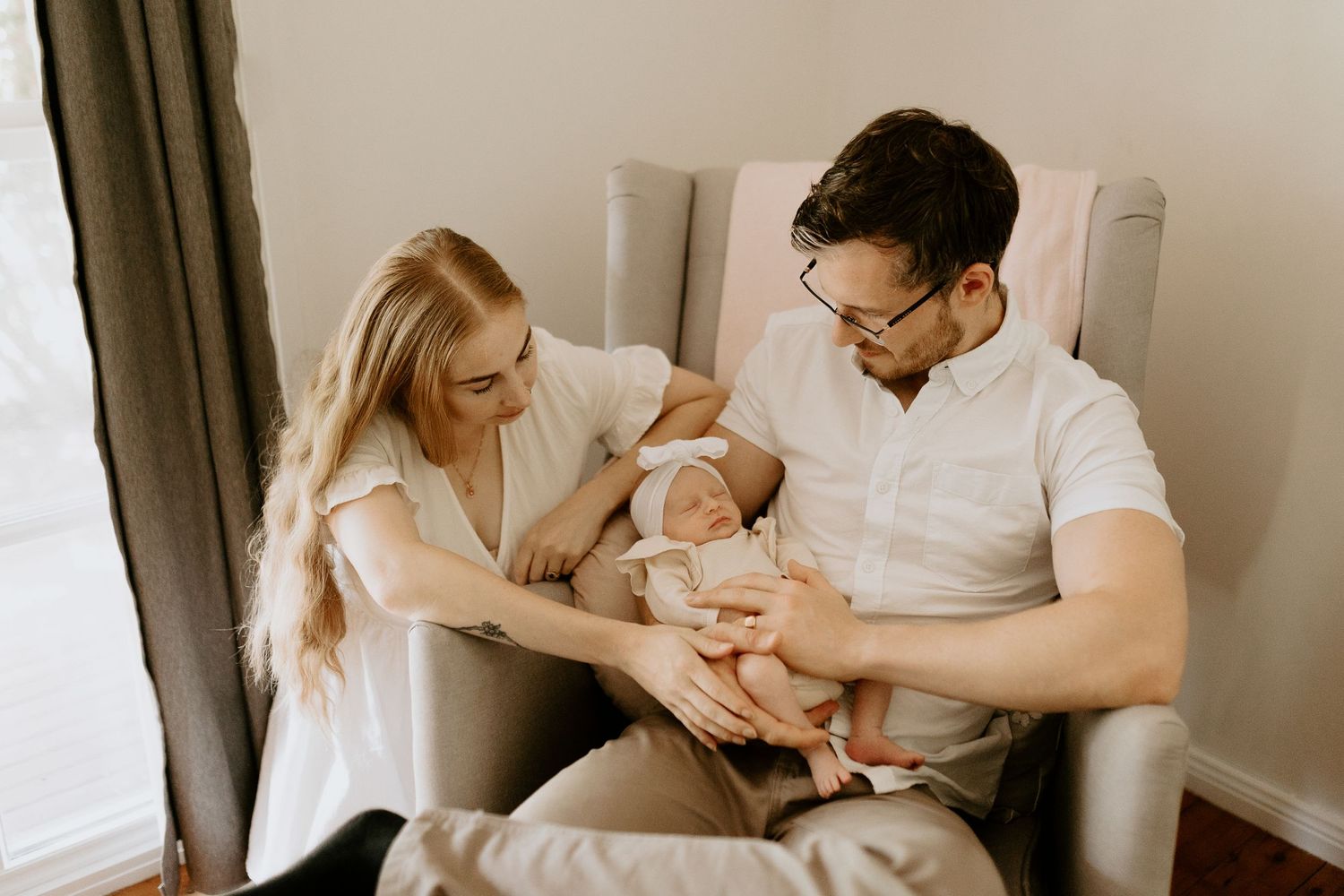 Parents lovingly embrace their newborn baby while sitting together by a bright window.