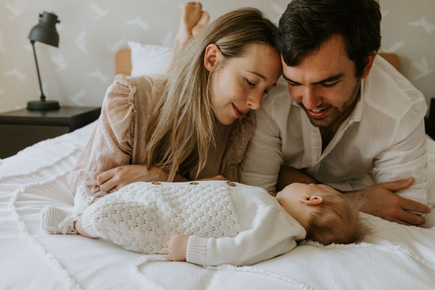 Family lying together on white bedding in soft natural light during intimate photo session.