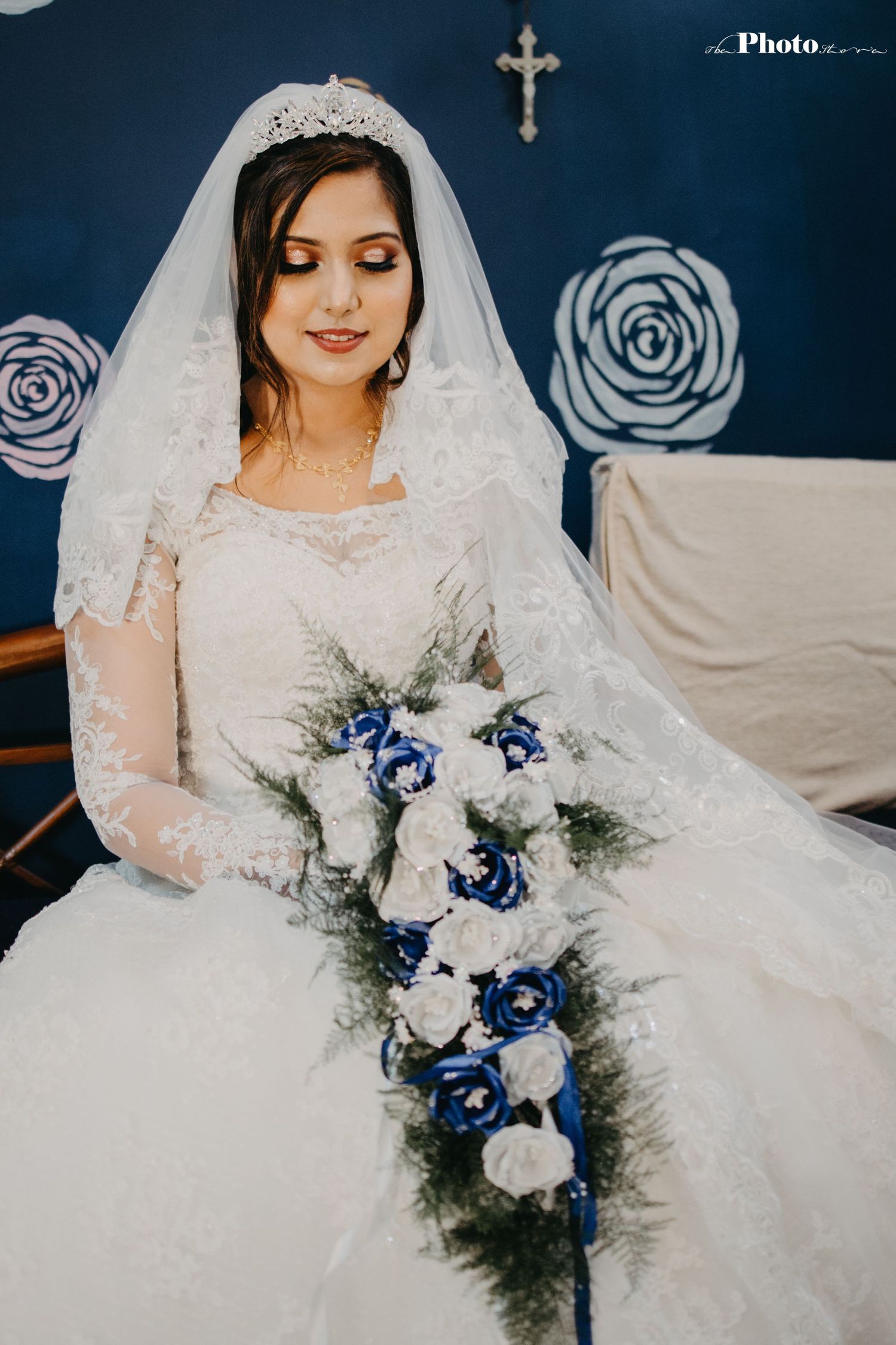 Bride in white lace dress holding blue and white bouquet against navy wall with rose patterns.