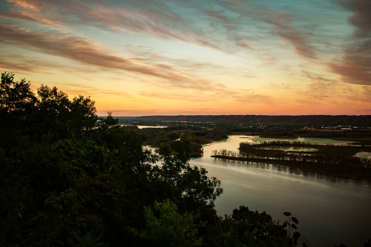 Dramatic sunset over winding river with silhouetted trees in foreground.