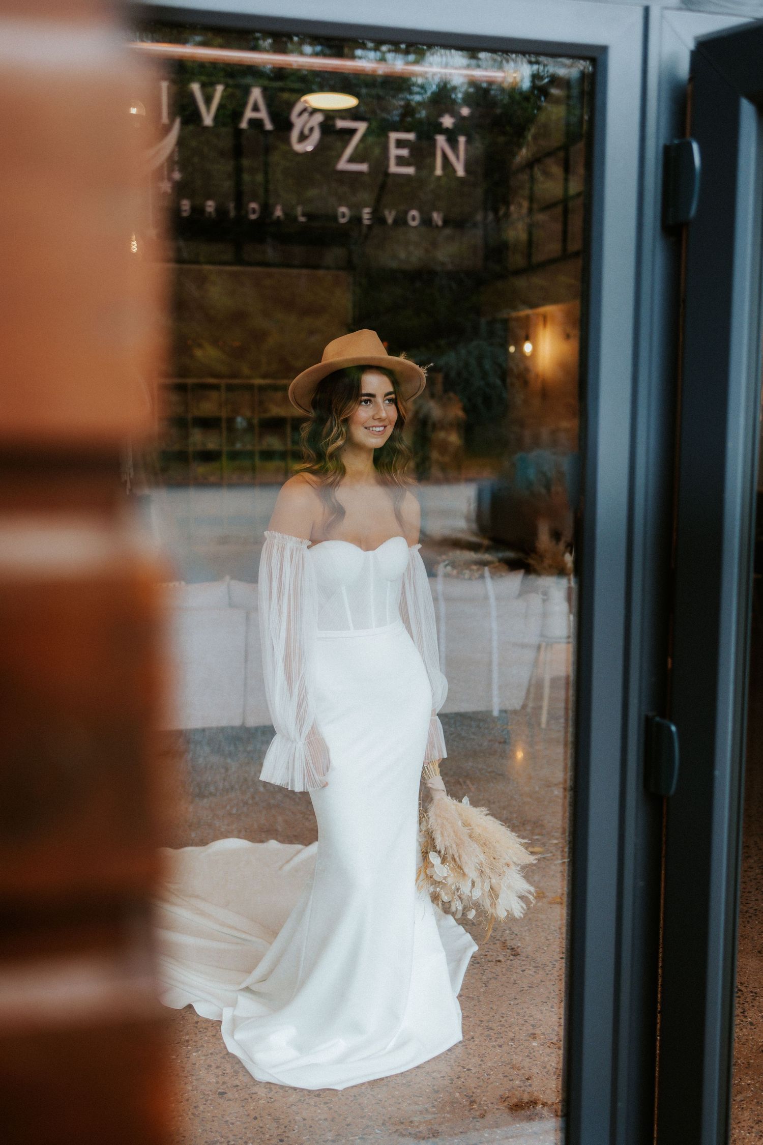 Bridal portrait through window reflection at Iva & Zen boutique featuring off-shoulder wedding gown.
