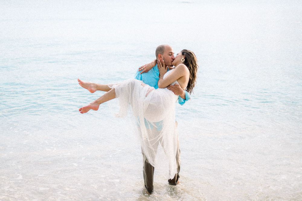 Romantic beach wedding photo with couple in white attire by ocean waters.