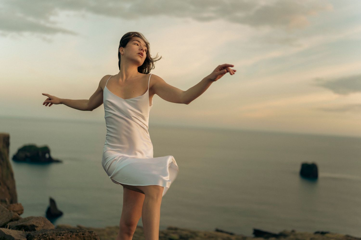A person in a white dress dances gracefully against a coastal sunset backdrop with rocks visible in the ocean.