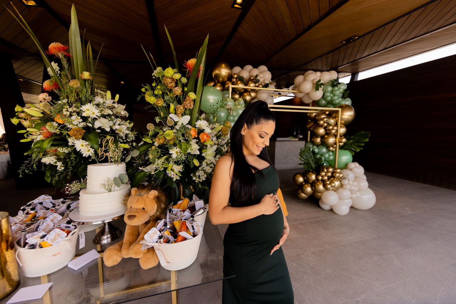 Pregnant woman in green dress stands near floral arrangements and balloon decor at a baby shower.
