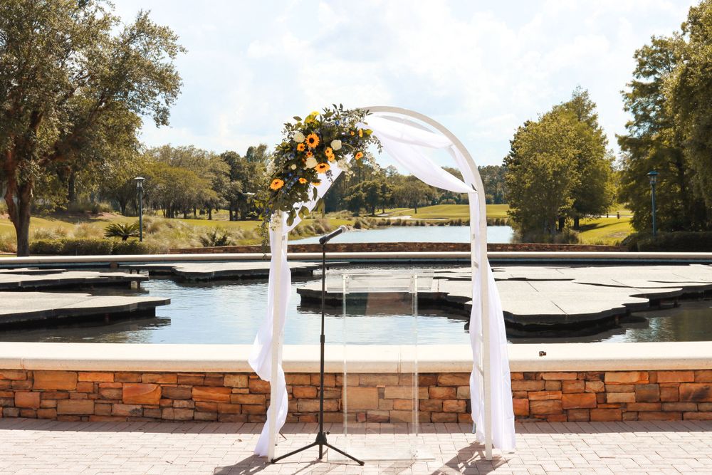 White draped wedding arch decorated with orange and yellow flowers stands on a brick patio.