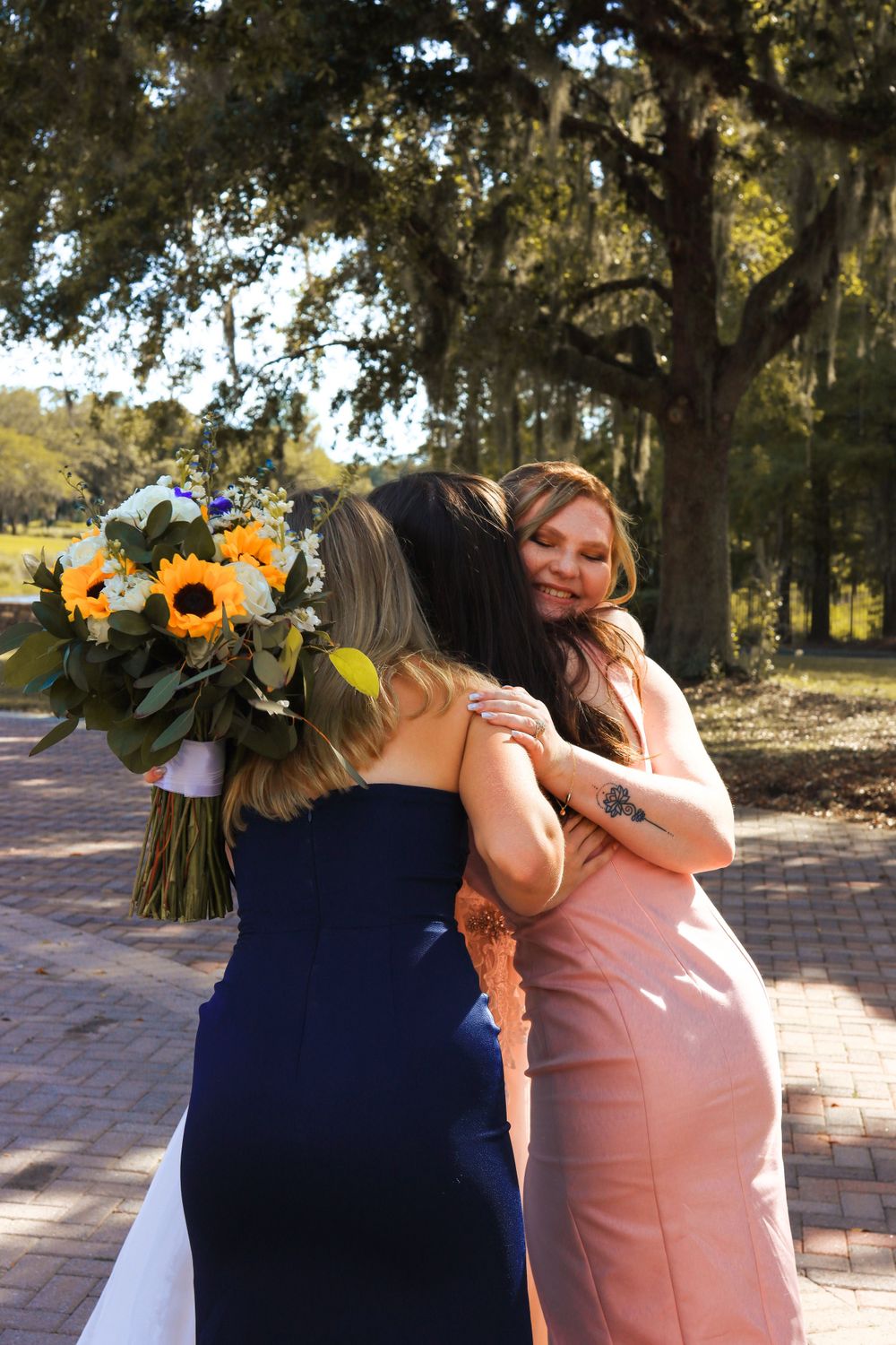 Two people embrace outdoors while one holds a bouquet of sunflowers on a brick path under oak trees.