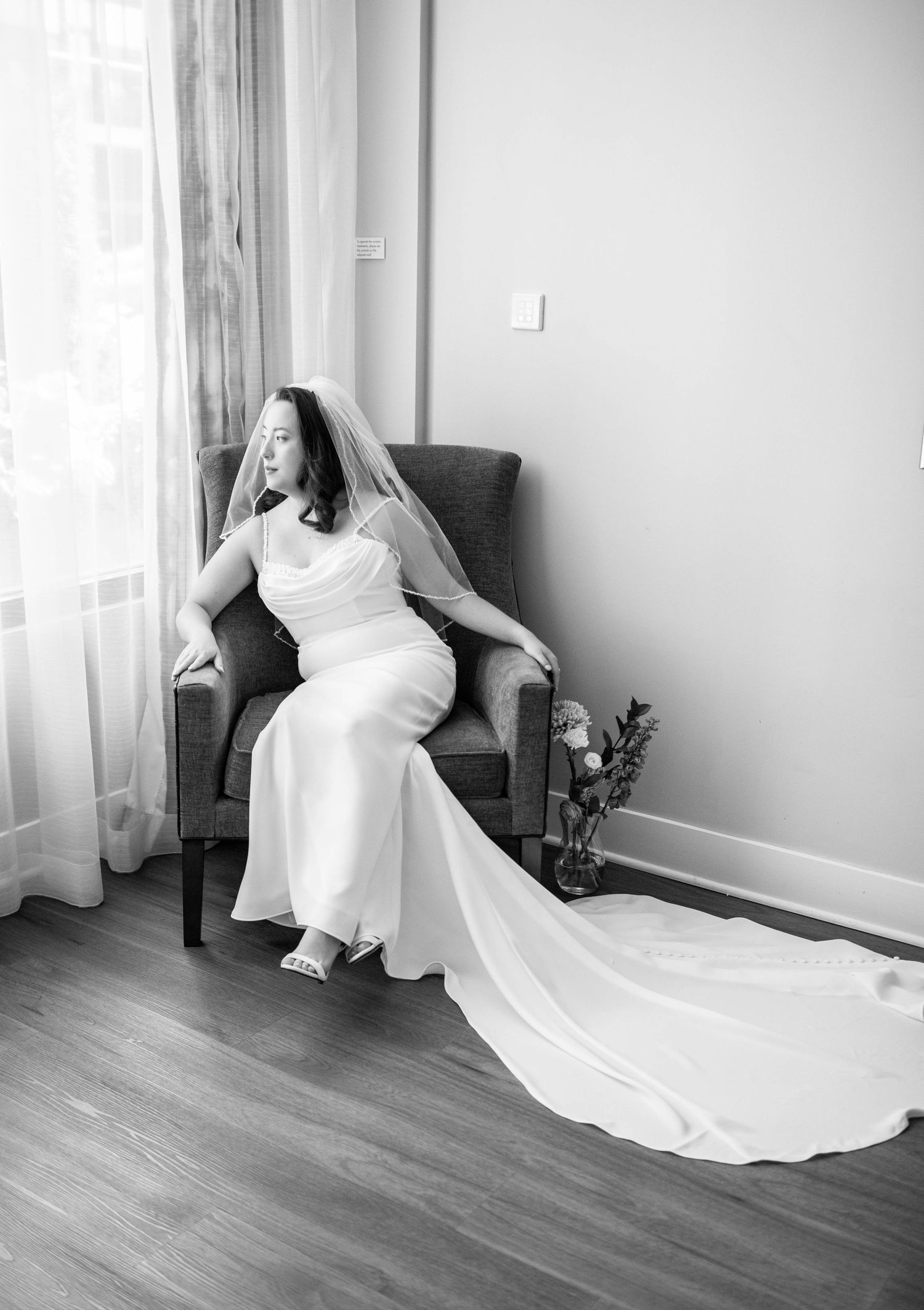 Bride in elegant white dress relaxes in grey armchair by window with flowing veil.