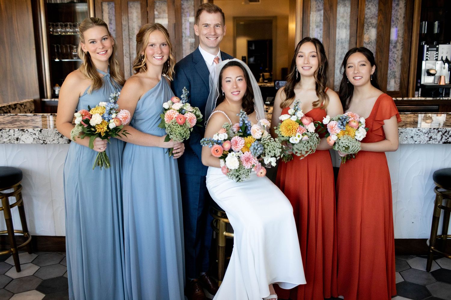 Wedding party posing at venue with bridesmaids in blue and rust-colored dresses.