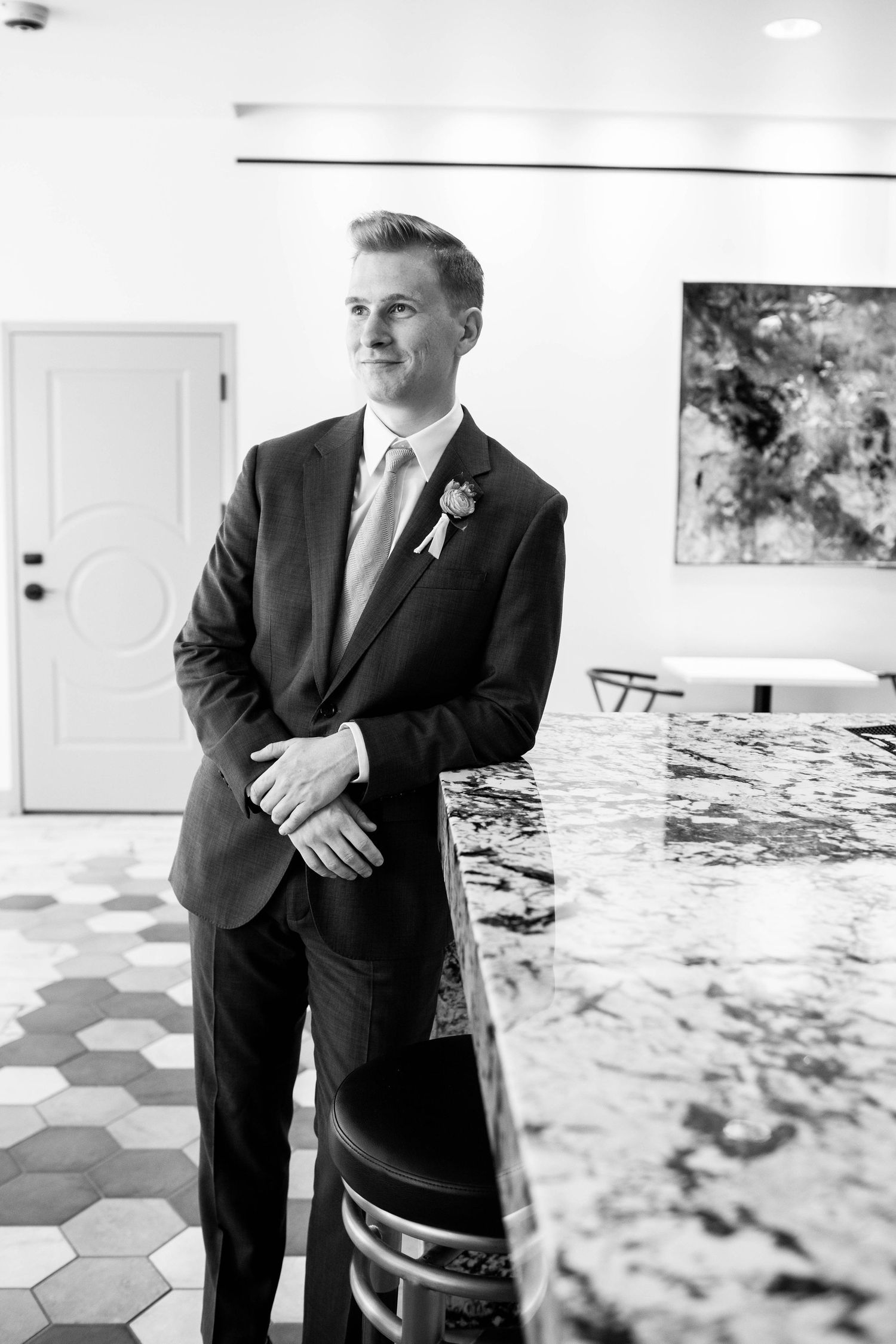 Groom in classic dark suit poses against geometric tile floor in black and white photo.