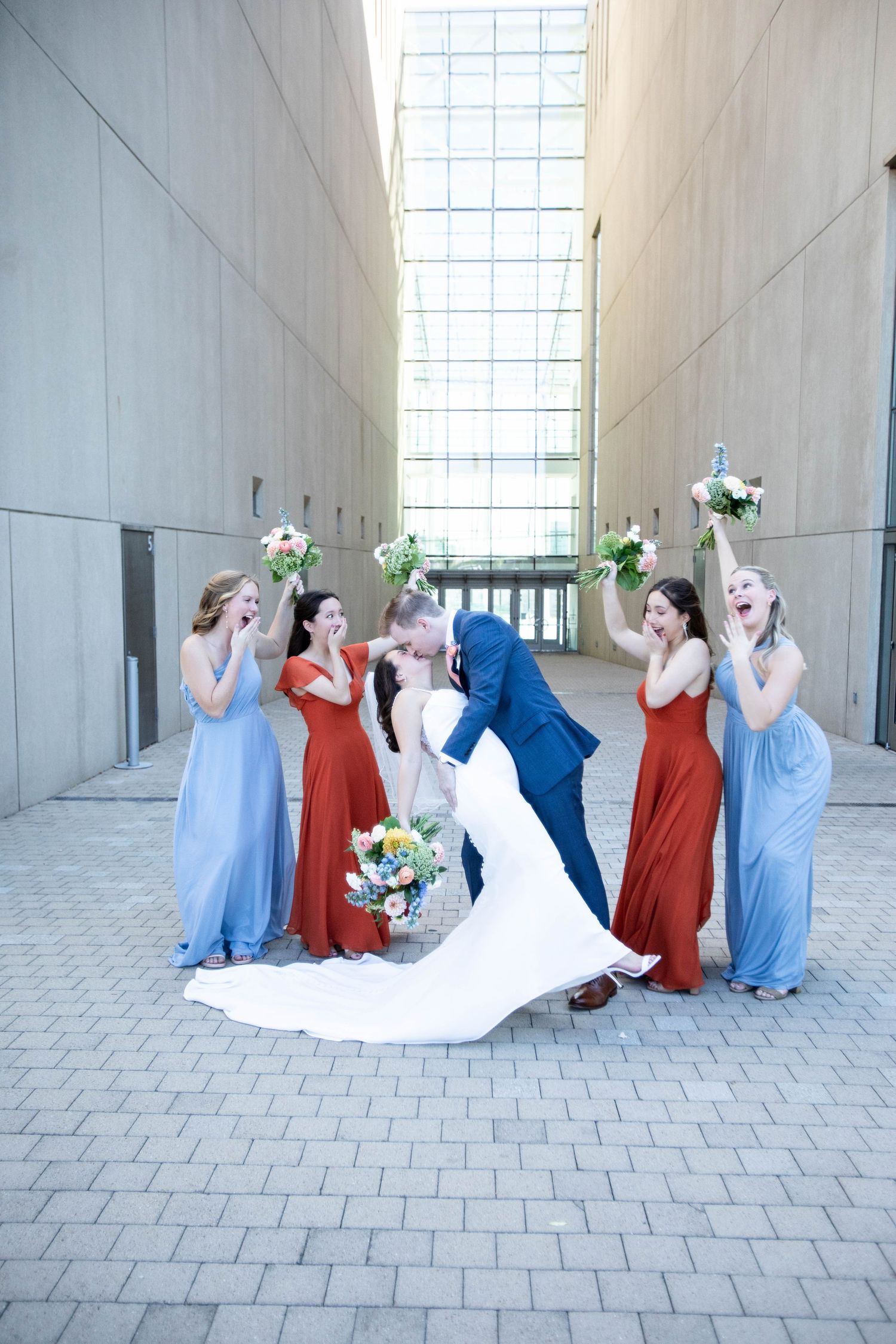 Fun bridal party photo with bridesmaids in red and blue dresses reacting to couple's kiss.