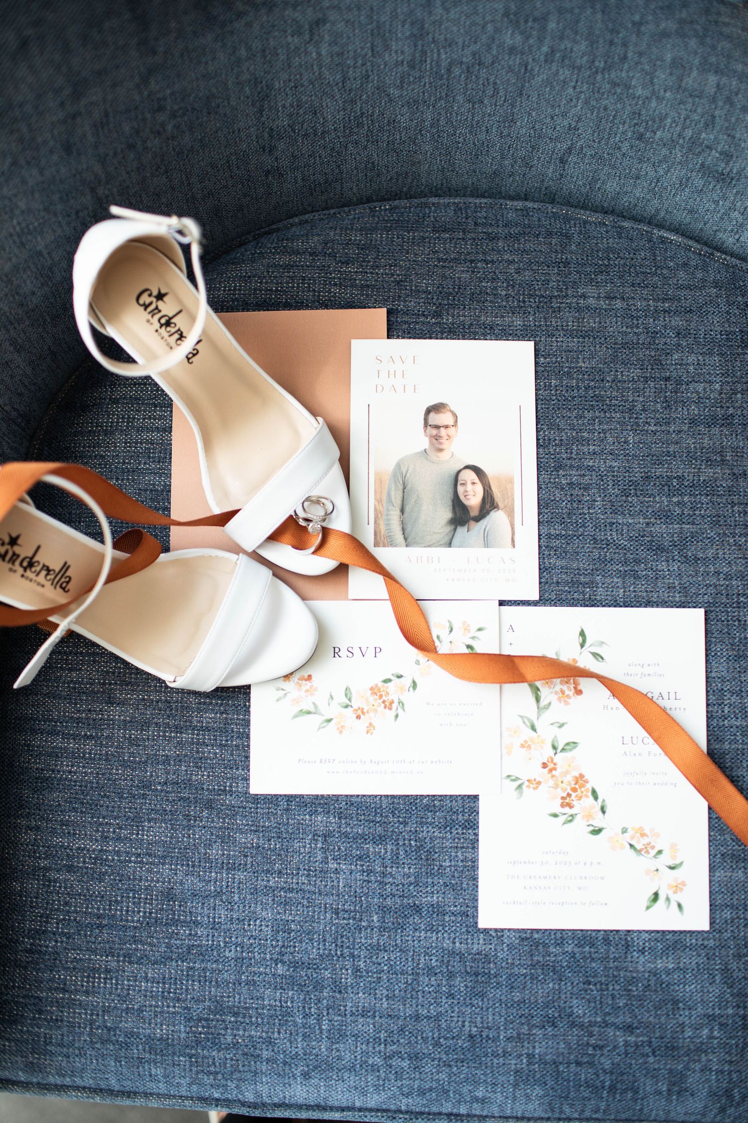 White wedding shoes and floral invitation cards arranged on a gray couch with an orange ribbon accent.