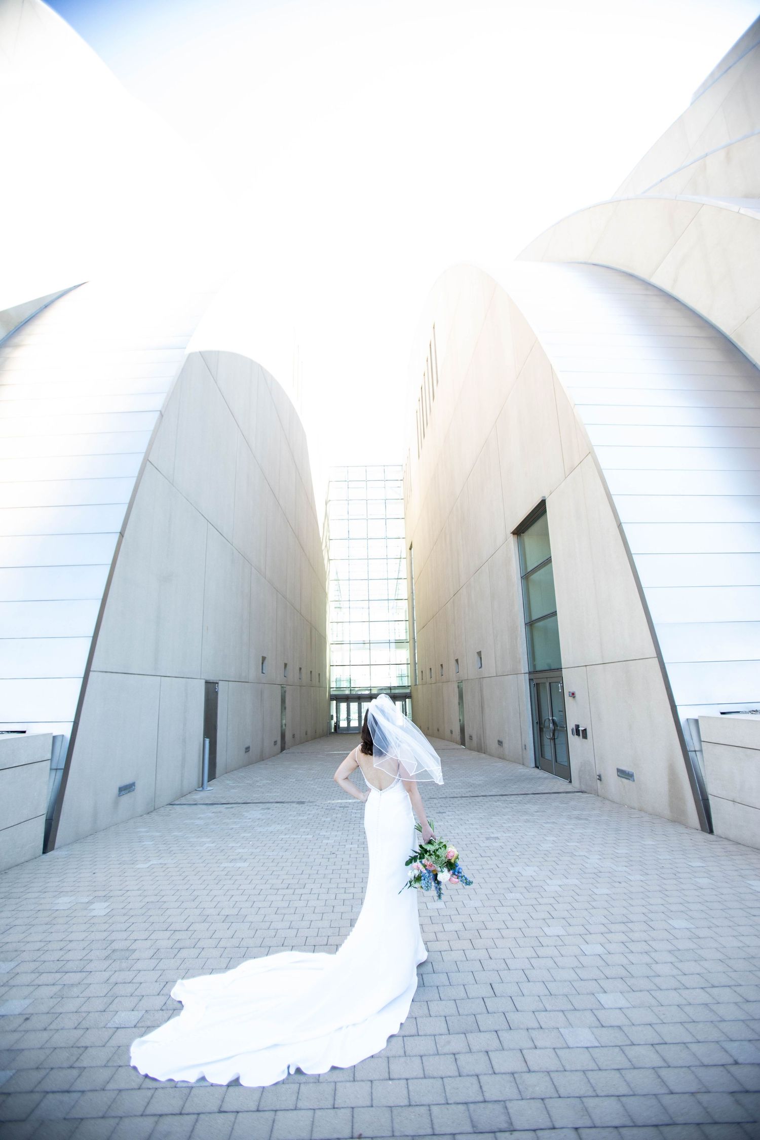 Bride in flowing white gown walks through modern architectural corridor holding a bouquet.