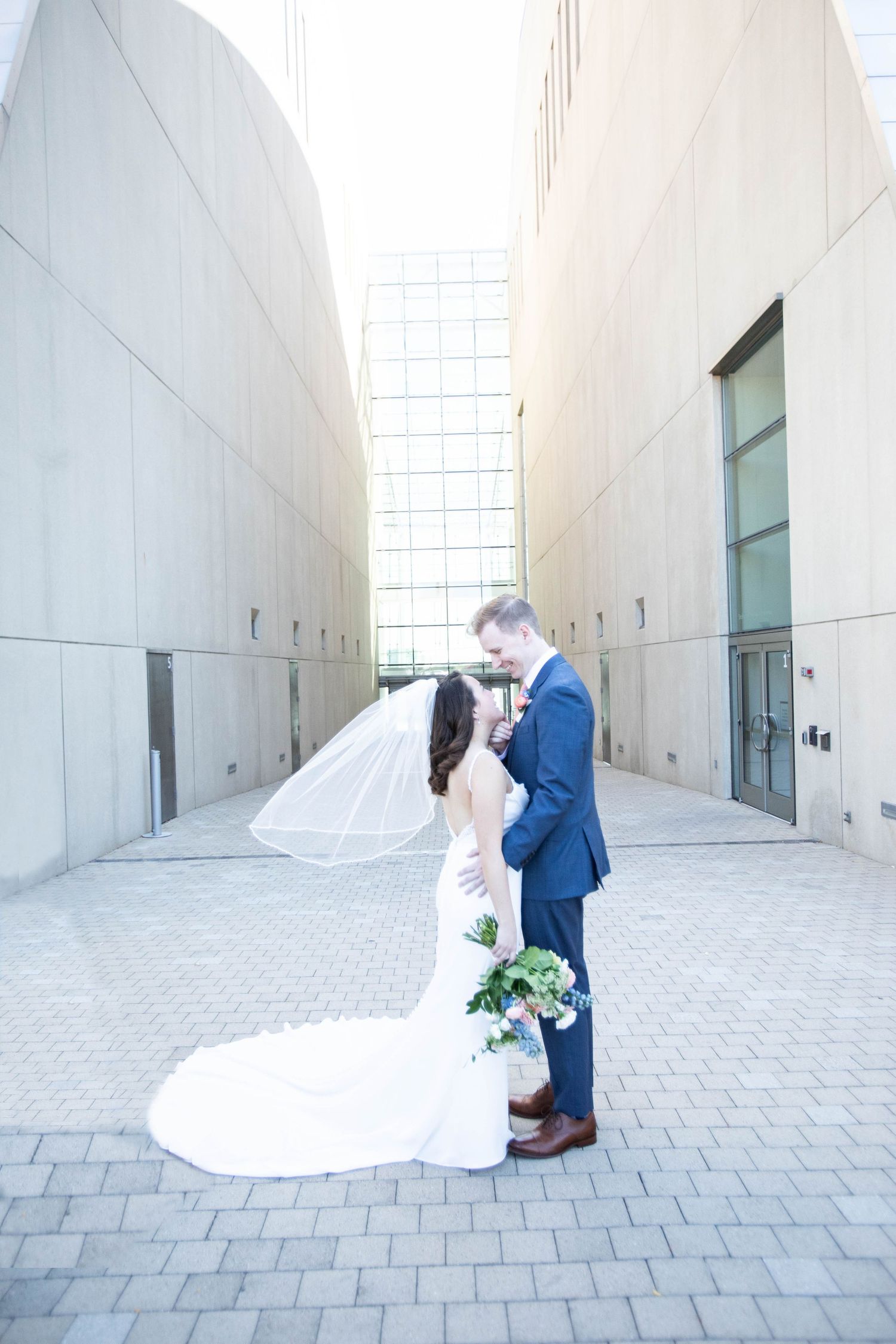 Couple in wedding attire share an intimate moment in an urban courtyard with dramatic white walls.