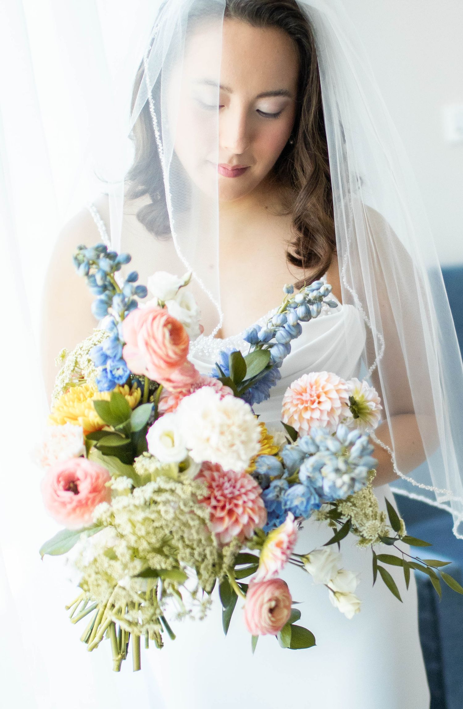 Wedding bouquet features pastel pink and blue flowers with delicate veil in foreground.