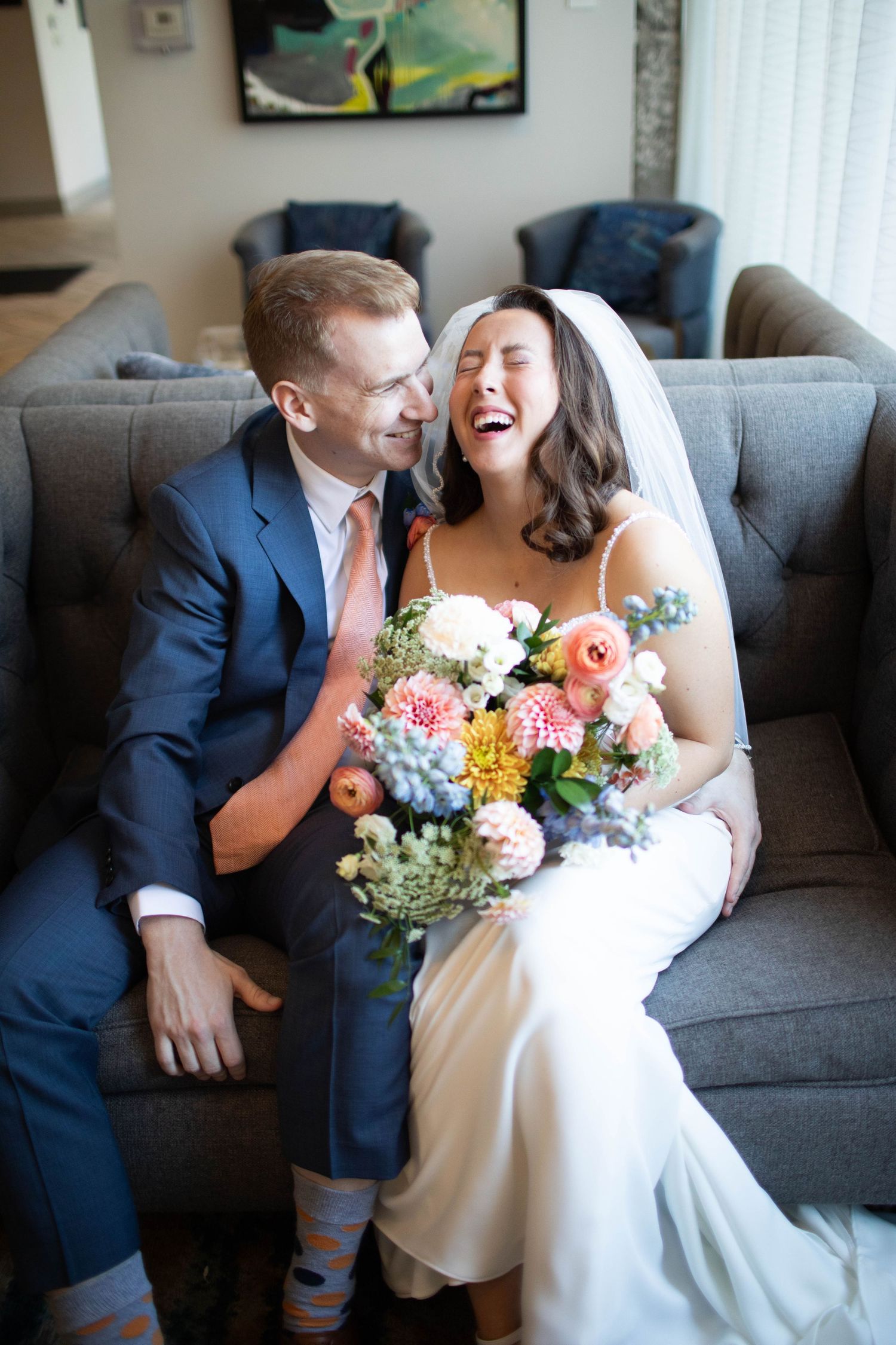 Joyful wedding moment on grey couch with colorful bouquet featuring roses and hydrangeas.