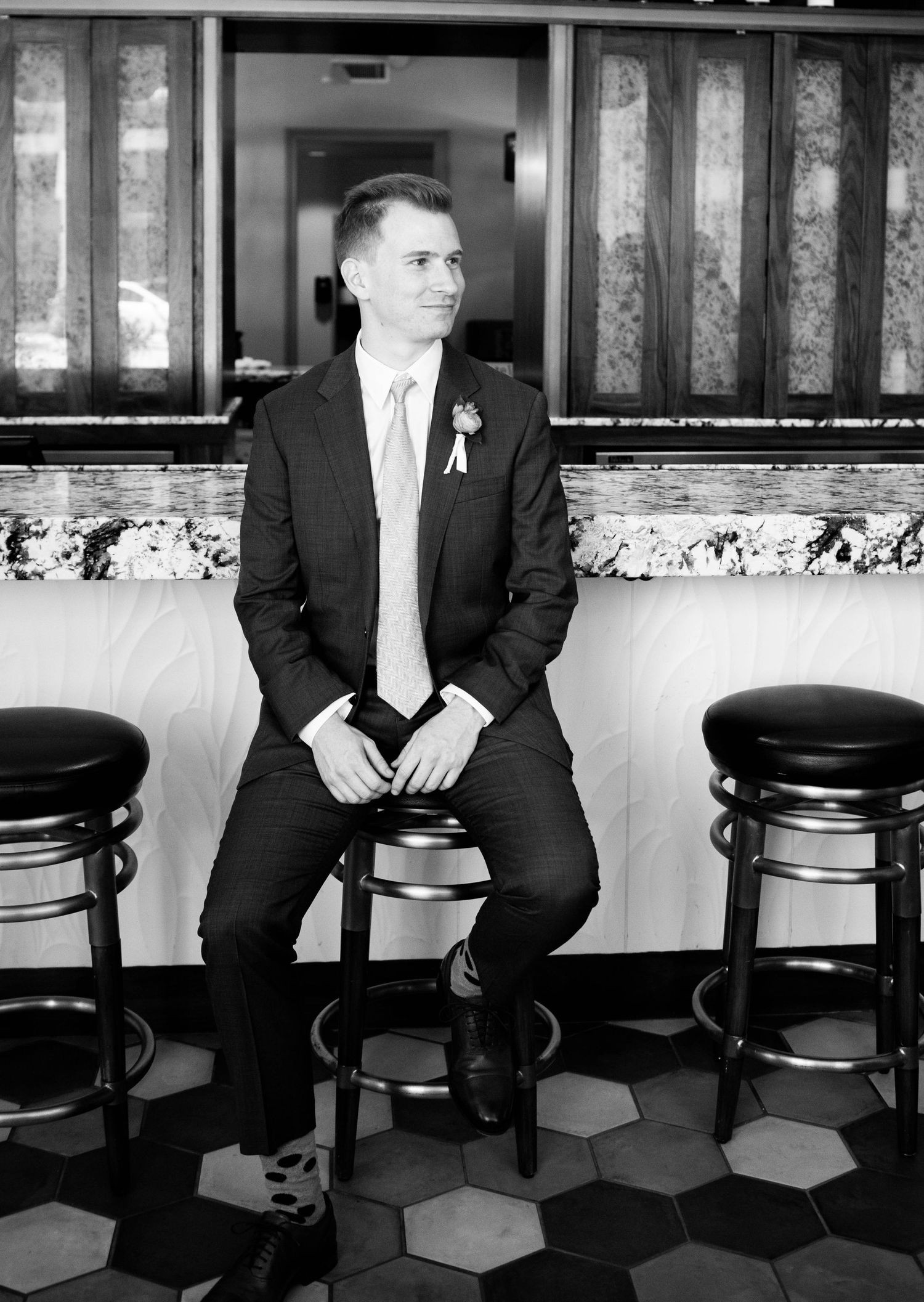 Groom in formal suit sits on bar stool in black and white photograph.