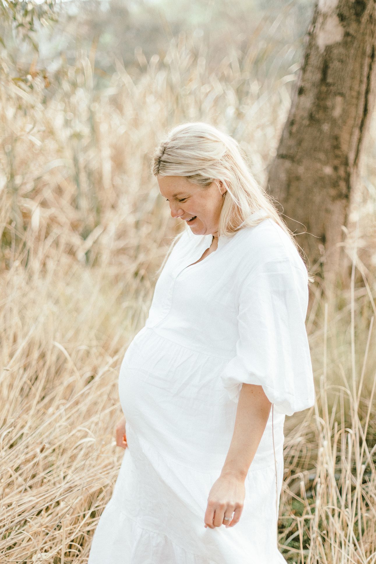 Expectant mother in flowing white dress poses in field of tall grass.