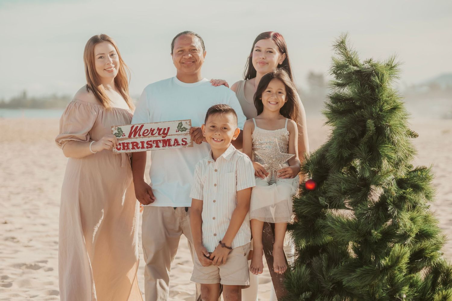 Family of five posing with Merry Christmas sign beside a decorated tree on a sandy beach.