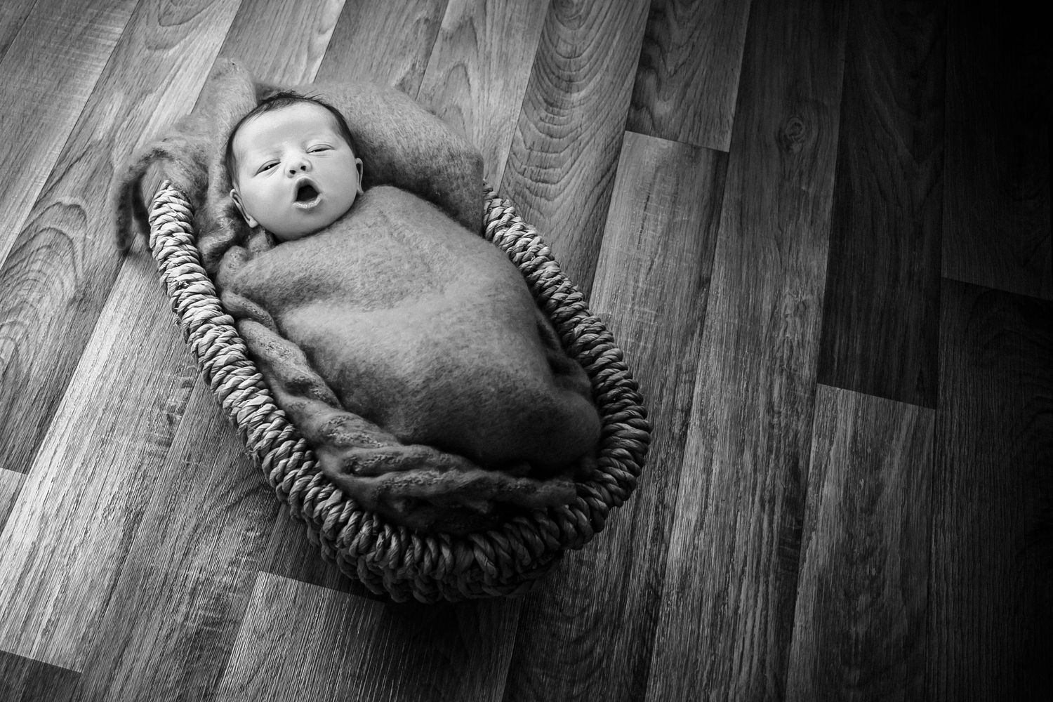 black and white photo of a baby wraped in a swollen blanke in a basket on a wooden floor with her pulling a funny face.