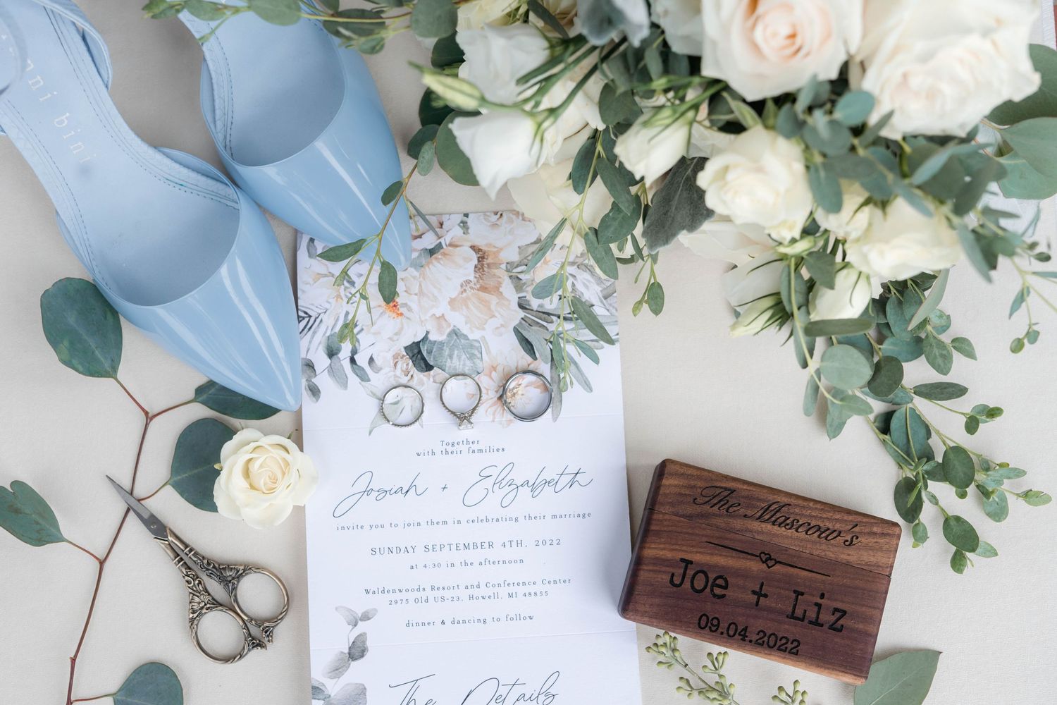 A wedding invitation and rings are displayed on a floral arrangement.