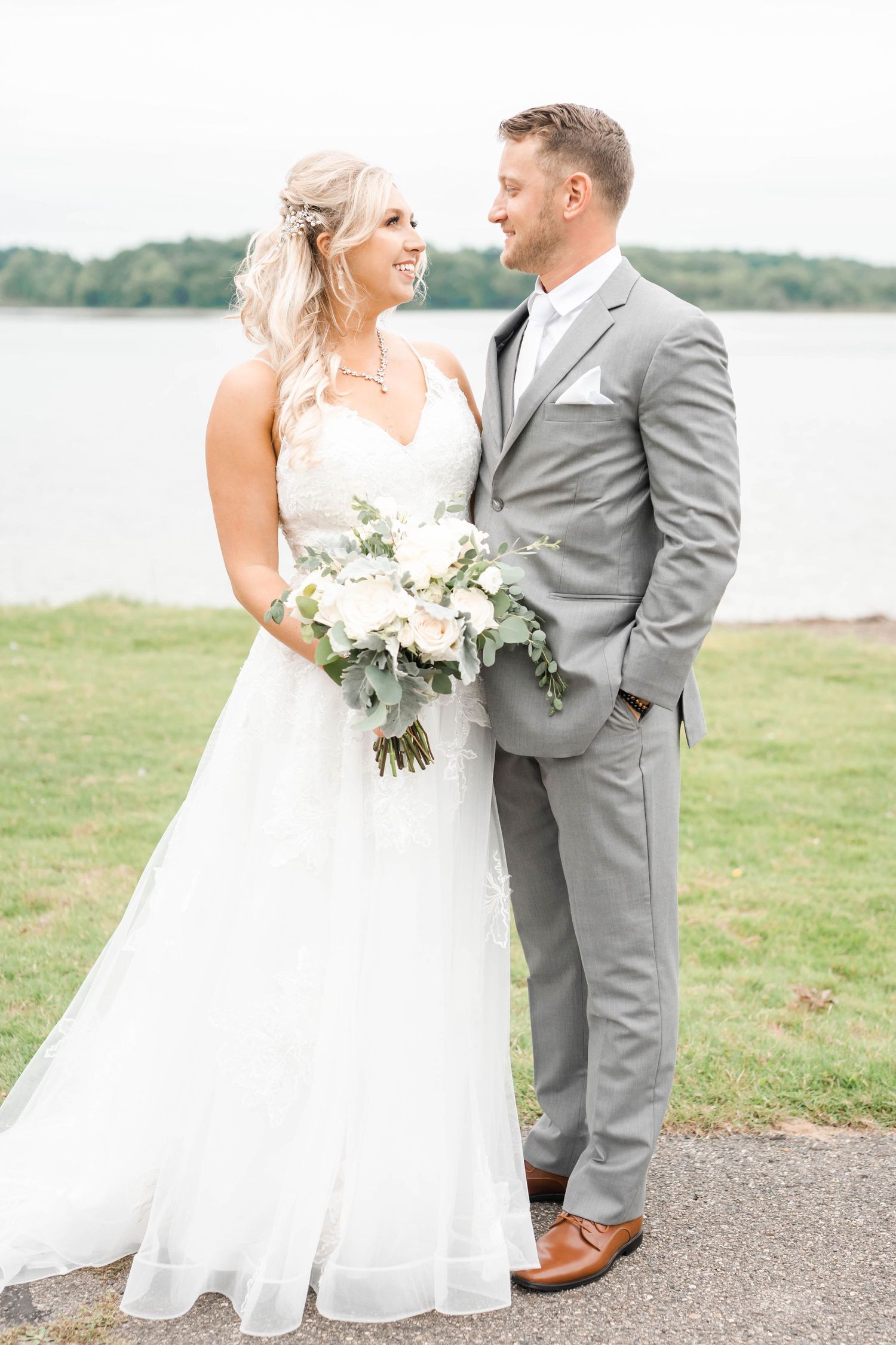 The image shows a bride and groom posing for a portrait on a grassy field near a body of water.