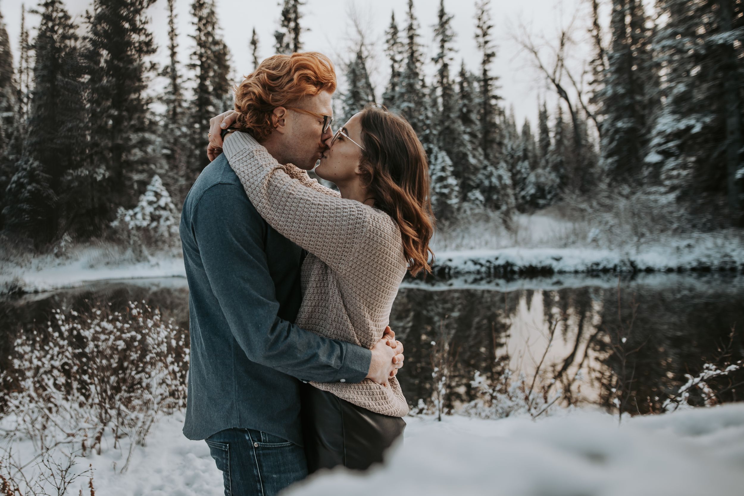 A couple shares a passionate kiss near a snowy lake surrounded by pine trees in winter.
